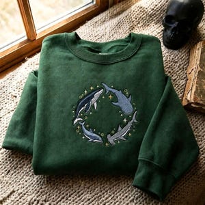 May include: A forest green crewneck sweatshirt featuring an embroidered circular design on the chest. The embroidery depicts four marine creatures, including a whale and sharks, arranged in a ring with small yellow stars and bubbles surrounding them.