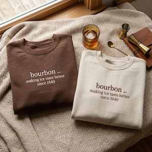 May include: A dark brown sweatshirt with white text and a cream sweatshirt with dark brown text, both featuring the embroidered phrase bourbon .. making ice taste better since 1840. The garments are displayed on a textured fabric surface.