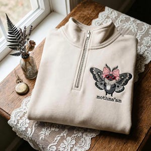 May include: A beige quarter-zip sweatshirt featuring an embroidered graphic of a moth wearing a pink bow and red sunglasses while holding a white cup. The text "mothma'am" is embroidered in black lettering below the moth graphic.