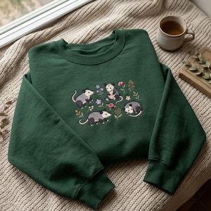 May include: A dark green crewneck sweatshirt featuring an embroidered design of four small opossums surrounded by colorful flowers and greenery. The embroidery includes a small pink button accent integrated into the floral pattern.