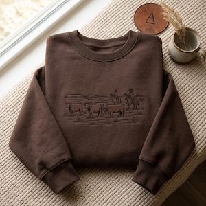 May include: A dark brown crewneck sweatshirt featuring a tonal embroidered graphic on the chest. The embroidery depicts a western scene with three cattle standing in a field and two figures on horseback in the background, all rendered in dark thread.