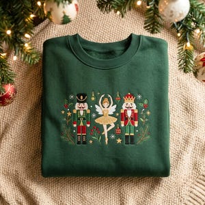 May include: A dark green crewneck sweatshirt featuring an embroidered design of two nutcrackers flanking a ballerina in a gold tutu. The scene is decorated with small ornaments, holly leaves, red berries, and gold stars arranged in a horizontal row.