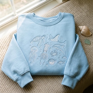 Op de afbeelding: Een lichtblauw sweatshirt met ronde hals en witte borduursels van diverse zeedieren, waaronder een zeeschildpad, pijlstaartrog, kwal, inktvis, krab, dolfijn, kogelvis en een kleine vis, allemaal omringd door kleine luchtbelletjes.