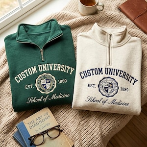 May include: A dark green and a cream-colored quarter-zip sweatshirt, each featuring the text CUSTOM UNIVERSITY, EST. 1889, and School of Medicine, along with a circular crest logo containing a book, a lamp, and a shield design on the chest.