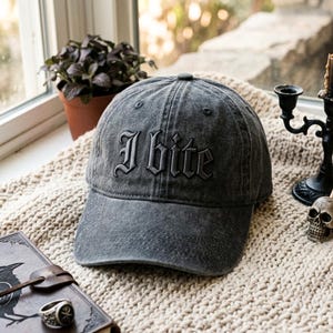 May include: A charcoal gray baseball cap with a distressed, washed-out finish. The front of the cap features the text "I bite" embroidered in a bold, black gothic-style font. The cap has a structured crown, a curved brim, and a button on top.