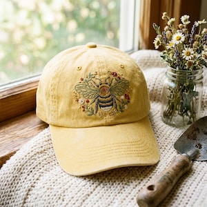 May include: A mustard yellow baseball cap featuring an embroidered design of a bumblebee surrounded by small red and white flowers and green leaves. The cap has a curved brim and a button on the top, with visible stitching details on the front panel.