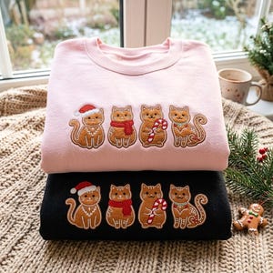 Op de afbeelding: Een lichtroze sweatshirt en een zwart sweatshirt op elkaar gestapeld, elk met een geborduurd ontwerp van vier bruine peperkoekkatten, één met een kerstmuts, één met een rode sjaal, één met een zuurstok en één met een groene halsband.