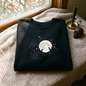 May include: A black crewneck sweatshirt featuring an embroidered design of three black cats surrounding a large, pale yellow full moon. The moon is accented with small white stars and dots, creating a celestial scene centered on the chest area.