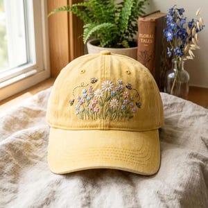 May include: A mustard yellow baseball cap featuring an embroidered design of white daisies, pink and blue wildflowers, and small black and yellow bees. The cap has a curved brim and a structured crown with stitched eyelets on the front panels.
