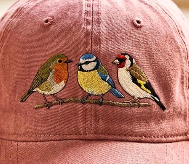 May include: A dusty rose baseball cap featuring an embroidered design of three small birds perched on a thin brown branch. The birds include a robin with an orange breast, a blue tit with yellow and blue feathers, and a goldfinch with red markings.