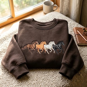 May include: A dark brown crewneck sweatshirt featuring an embroidered design of four galloping horses in a row. The horses are depicted in shades of dark brown, light tan, white, and black, showcasing detailed manes and tails on the fabric.