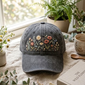 May include: A charcoal gray baseball cap featuring detailed embroidery of various wildflowers across the front panel. The design includes small daisies, poppies, and leafy stems in shades of white, pink, orange, and green stitched onto the fabric.