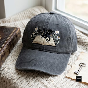 Puede incluir: Una gorra de béisbol gris carbón con un bordado de un pequeño dragón negro posado sobre un libro abierto, rodeado de margaritas blancas con tallos y hojas verdes. La gorra tiene una visera curva y un diseño estructurado de seis paneles.