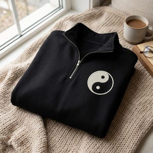 Könnte beinhalten: Ein schwarzes Sweatshirt mit kurzem Reißverschluss und einem kreisförmigen Yin-Yang-Aufnäher auf der linken Brust. Das Kleidungsstück verfügt über einen silberfarbenen Metallreißverschluss am Kragen und besteht aus einfarbigem Stoff.