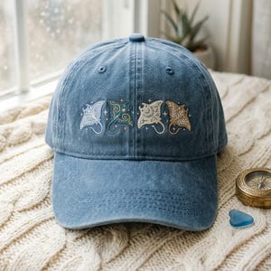Pode incluir: Um boné de beisebol de ganga azul com um design bordado de quatro raias manta em vários tons de branco, azul-petróleo e bege, acentuado com pequenos motivos de estrelas nos painéis frontais do boné acima da aba curva.