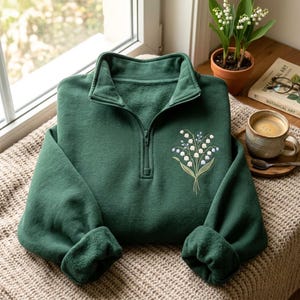 May include: A dark green quarter-zip sweatshirt featuring an embroidered bouquet of white lily of the valley flowers with green stems and leaves on the upper left chest. The garment has a high collar, long sleeves, and a soft fleece-like texture.