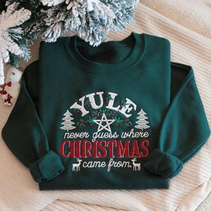 Może przedstawiać: Ciemnozielona bluza z okrągłym dekoltem i białym oraz czerwonym haftem. Tekst brzmi YULE never guess where CHRISTMAS came from. Wzór zawiera centralny pentagram, dwie sosny, jagody ostrokrzewu oraz dwie małe sylwetki jeleni.