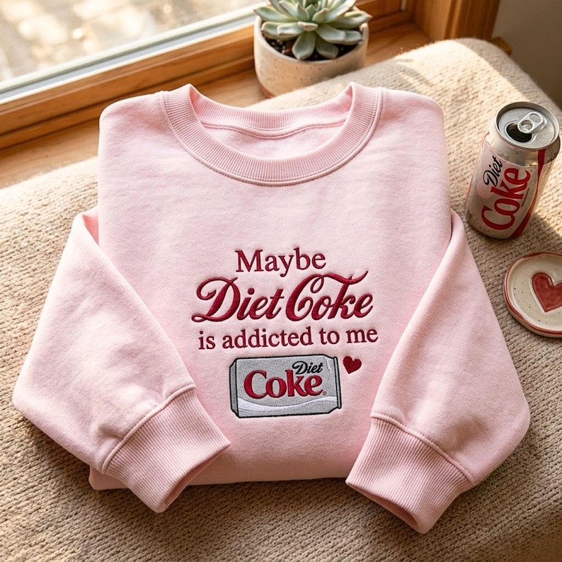 Pu&ograve; includere: Una felpa girocollo rosa chiaro con testo ricamato in rosso scuro che recita "Maybe Diet Coke is addicted to me" sopra una piccola grafica ricamata di una lattina di Diet Coke argentata e una minuscola icona a forma di cuore rosso.