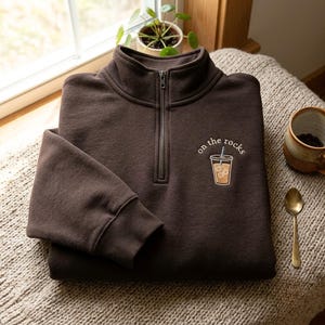 Könnte beinhalten: Ein dunkelbraunes Sweatshirt mit kurzem Reißverschluss und einer Stickerei auf der linken Brust. Das Motiv zeigt einen durchsichtigen Kunststoffbecher mit einem Kaltgetränk, einem Strohhalm und dem weißen Schriftzug "on the rocks" darüber.