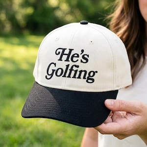 May include: A baseball cap with an off-white crown and a solid black brim. The front of the crown features the text He's Golfing embroidered in black, cursive-style lettering. The cap includes a small black button at the top and stitched eyelets.