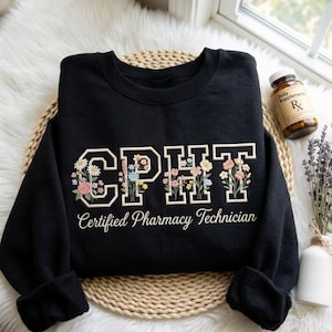 May include: A black crewneck sweatshirt featuring the embroidered letters CPHT in a collegiate font, with colorful floral designs integrated into each letter. Below the letters, the text Certified Pharmacy Technician is embroidered in white.