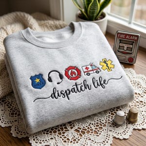 May include: A light gray crewneck sweatshirt featuring embroidered icons of a blue police badge, black headset, red fire emblem, ambulance, and yellow star of life. Below the icons, the text "dispatch life" is embroidered in black script.