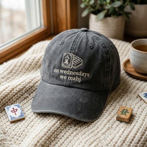 May include: A charcoal gray baseball cap featuring an embroidered graphic of two mahjong tiles and the text "on wednesdays we mahj" in a light cream color. The cap has a structured crown, a curved brim, and stitched eyelets for ventilation.