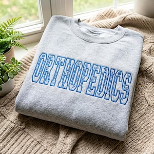 Könnte beinhalten: Ein hellgraues Sweatshirt mit Rundhalsausschnitt und dem Wort ORTHOPEDICS, das in blauen Blockbuchstaben gestickt ist. Die Buchstaben sind mit blauem Garn umrandet und mit einem zarten, sich wiederholenden Blumenrankenmuster gefüllt.