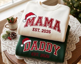 May include: A cream-colored sweatshirt with the red text MAMA and EST. 2026, and a dark green sweatshirt with the red text DADDY and EST. 2026. Both feature a red and white Santa hat graphic above the primary text in a collegiate font.