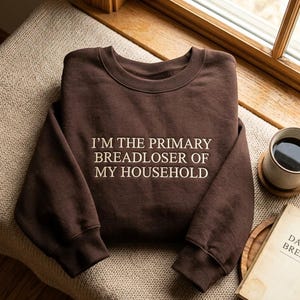 May include: A dark brown crewneck sweatshirt featuring the white embroidered text I'M THE PRIMARY BREADLOSER OF MY HOUSEHOLD centered on the chest. The garment has long sleeves with ribbed cuffs and a ribbed neckline, presented in a simple design.