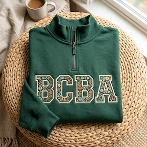May include: A dark green quarter-zip sweatshirt featuring the letters BCBA embroidered on the front. Each letter is filled with a detailed floral pattern in shades of orange, blue, and cream, outlined in a cream-coloured thread.