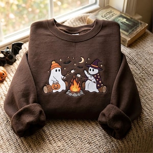Könnte beinhalten: Ein dunkelbraunes Sweatshirt mit Rundhalsausschnitt und einer Stickerei von zwei weißen Geistern, die auf Baumstämmen an einem Lagerfeuer sitzen. Ein Geist trägt eine orangefarbene Mütze und einen Kürbiseimer, der andere einen Hexenhut und Umhang.
