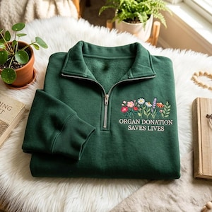 Puede incluir: Una sudadera verde oscuro con cremallera de un cuarto y un diseño bordado de flores silvestres coloridas sobre el texto "ORGAN DONATION SAVES LIVES" en letras mayúsculas blancas. La prenda tiene una cremallera metálica plateada.