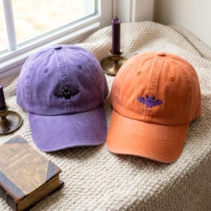 Può includere: Un cappellino da baseball viola con un pipistrello nero ricamato e un cappellino arancione con un pipistrello viola ricamato. Entrambi i cappellini presentano una struttura a sei pannelli con visiera curva e un bottone sulla parte superiore.
