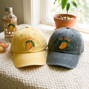 May include: Two baseball caps, one yellow and one charcoal gray, each featuring an embroidered yellow mango with green leaves and the text "let that" stitched in orange thread above the fruit design on the front panel of the hat.