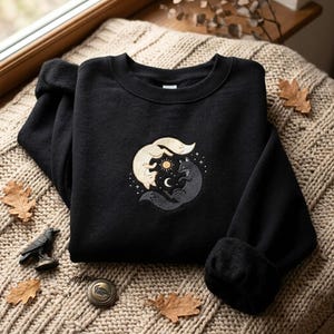 Könnte beinhalten: Ein schwarzes Sweatshirt mit Rundhalsausschnitt und einer Stickerei von zwei Füchsen in kreisförmiger Anordnung. Ein Fuchs ist hellcremefarben, der andere dunkelgrau. Dazwischen befinden sich eine kleine gelbe Sonne, ein weißer Mond und Sterne.