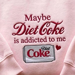 Pu&ograve; includere: Una felpa rosa chiaro con testo ricamato in rosso scuro che recita "Maybe Diet Coke is addicted to me" sopra un grafico ricamato di una lattina di Diet Coke argento e rossa con una piccola icona a forma di cuore rosso a destra.