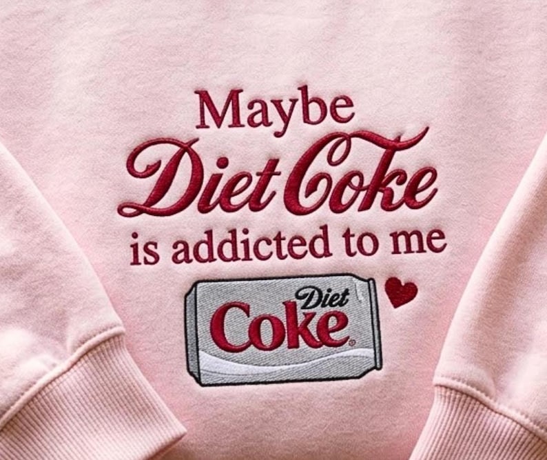 Pu&ograve; includere: Una felpa rosa chiaro con testo ricamato in rosso scuro che recita "Maybe Diet Coke is addicted to me" sopra un grafico ricamato di una lattina di Diet Coke argento e rossa con una piccola icona a forma di cuore rosso a destra.