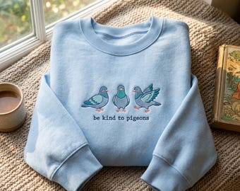 May include: A light blue crewneck sweatshirt featuring an embroidered design of three pigeons in shades of blue, teal, and grey. Below the birds, the text "be kind to pigeons" is stitched in a dark blue font. The garment has ribbed cuffs and collar.