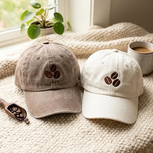 May include: Two baseball caps, one in a faded brown and one in off-white, each featuring an embroidered design of three dark brown coffee beans centred on the front panel. Both caps have a curved brim and stitched eyelets on the crown.