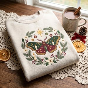 May include: A cream-colored crewneck sweatshirt featuring an embroidered design of a moth centered within a wreath of pine branches, white flowers, red berries, small ornaments, stars, and lit candles in shades of green, red, and gold.