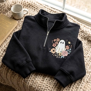 Peut inclure: Un sweat-shirt noir à fermeture éclair quart de tour avec une broderie sur la poitrine gauche. Le motif représente un fantôme blanc entouré d'une couronne florale colorée composée de fleurs roses, orange, bleues et de feuilles vertes.