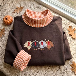 May include: A dark brown long-sleeve sweatshirt featuring a mock neck collar and sleeve cuffs in an orange and white gingham pattern. The center of the chest displays an embroidered design of four turkeys in different colors with small stars.