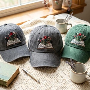 May include: Three baseball caps in grey and green featuring an embroidered design of an open book with colourful flowers blooming from the pages. The caps have a structured crown, a curved brim, and eyelets for ventilation on the top panels.