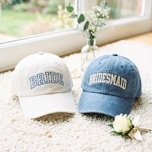 May include: A white baseball cap with the word BRIDE embroidered in blue and a blue denim baseball cap with the word BRIDESMAID embroidered in white. Both caps feature a curved brim, a button on top, and stitched eyelets for ventilation.