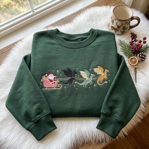 Pode incluir: Uma camisola de gola redonda verde-escura com um design bordado do Pai Natal num trenó cor-de-rosa puxado por três dragões em preto, verde-claro e dourado. O bordado inclui pequenas estrelas douradas espalhadas pelas figuras.