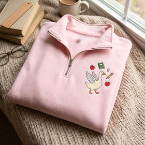 May include: A light pink quarter-zip sweatshirt featuring an embroidered design on the left chest. The embroidery depicts a white goose with spread wings surrounded by a green book, a yellow pencil, a paperclip, and two red apples.