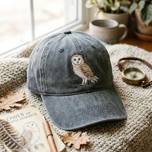Puede incluir: Una gorra de béisbol gris carbón con una lechuza común bordada en el panel frontal. La gorra tiene un acabado de tela desgastada, una visera curva y ojales cosidos en la corona, mostrando un diseño detallado de ave en blanco y marrón.