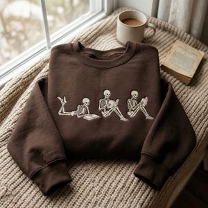 Op de afbeelding: Een donkerbruine sweater met ronde hals en een crèmekleurige grafische print van drie skeletten in verschillende houdingen, die elk een boek lezen. Het eerste skelet ligt op de buik, het tweede zit en het derde zit rechtop.