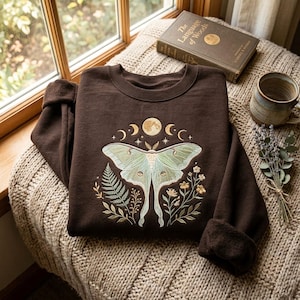 May include: A dark brown crewneck sweatshirt featuring an embroidered design of a large pale green moth centered above botanical elements including a fern and small flowers, with a moon phase cycle and stars stitched in gold thread above the moth.